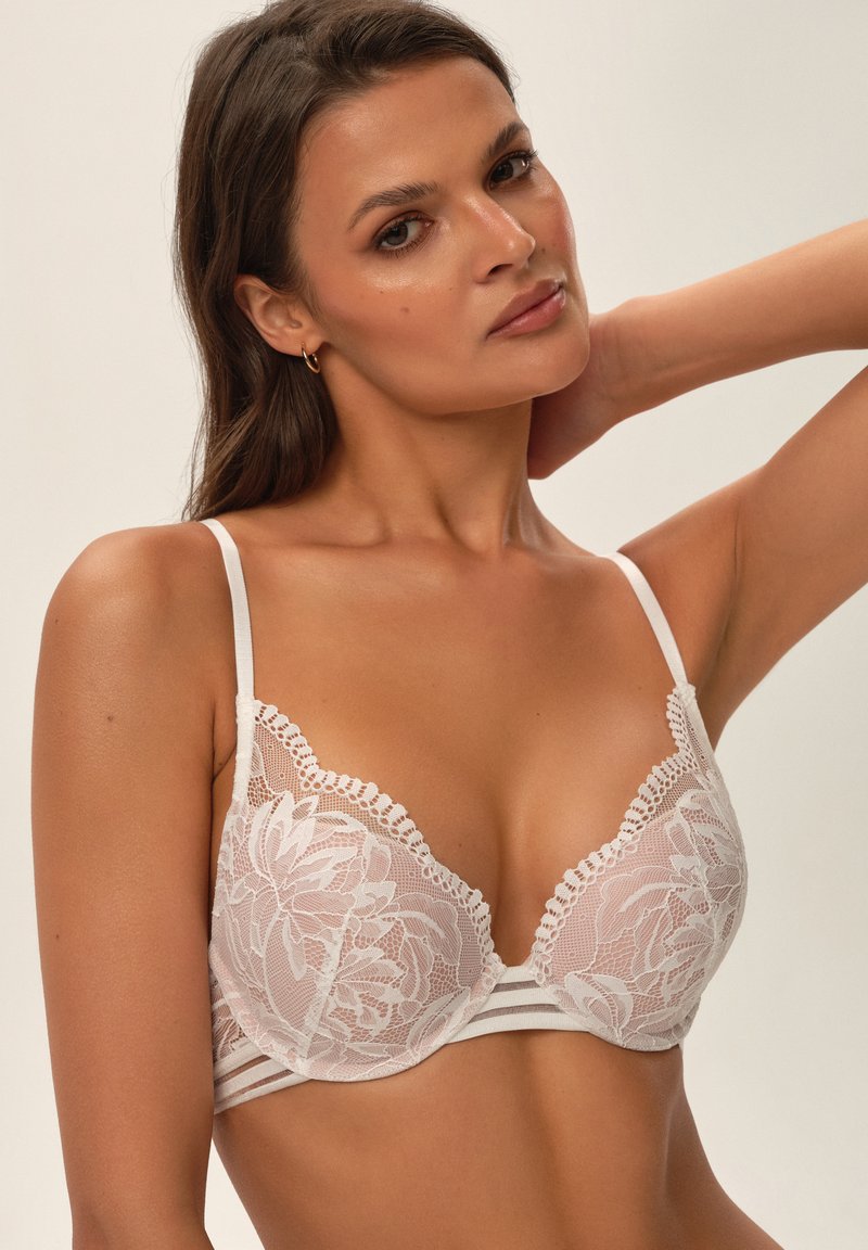 Lace bralette in light pink with floral pattern, featuring adjustable straps and a band with horizontal stripes. Textured lace trim at edges.