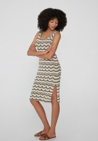 Strickkleid mit einem Zickzackmuster in erdigen Tönen von Grün, Beige und Weiß. Mit einem seitlichen Schlitz und einem Rundhalsausschnitt. Getragen mit flachen Sandalen.