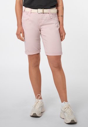 Persoon die lichtroze opgerolde korte broek draagt, een crèmekleurige geweven riem, witte sneakers en een zwart topje, staande op een effen lichte achtergrond.