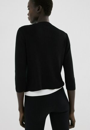 Mujer de espaldas que lleva una blusa de manga larga negra sobre una camisa blanca y pantalones negros, sobre un fondo claro y sencillo.