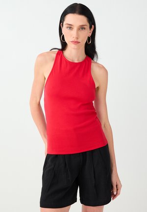Femme aux cheveux foncés portant des boucles d'oreilles anneaux dorées, un haut rouge sans manches et un short noir, debout devant un fond clair.
