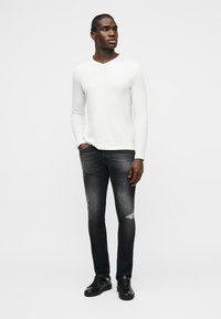 Άνδρας που φοράει λευκό πουκάμισο με μακριά μανίκια, μαύρα ξεβαμμένα τζιν με φθαρμένες λεπτομέρειες και μαύρα αθλητικά παπούτσια, στέκεται με το ένα χέρι στην τσέπη.