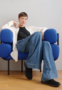 Blå polstret stol med avrundede former, sorte ben; person iført brede denimbukser og en sort top, siddende afslappet.