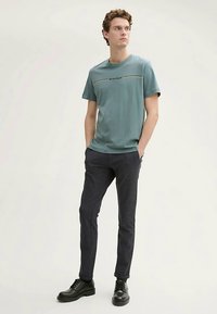 Světle tyrkysové bavlněné tričko s krátkými rukávy a vodorovným černým pruhem, doplněné tmavými texturovanými úzkými kalhotami a černými botami.