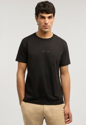 Giovane uomo in piedi con le mani nelle tasche dei pantaloni beige, indossando una t-shirt nera Trussardi su uno sfondo chiaro e uniforme.