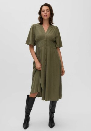 Woman wearing a knee-length olive green buttoned dress with short sleeves and black knee-high boots standing against a plain background.