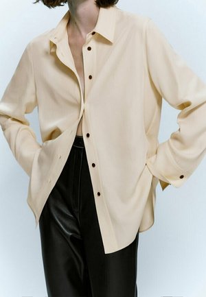 Osoba ubrana w oversize'ową beżową koszulę zapinaną na ciemne guziki, w połączeniu z czarnymi, wysokim stanem spodniami ze skóry, ręce w kieszeniach.