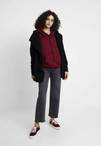 Un cardigan noir texturé par-dessus un sweat à capuche bordeaux, associé à un jean large gris et des baskets rouges à imprimé léopard.