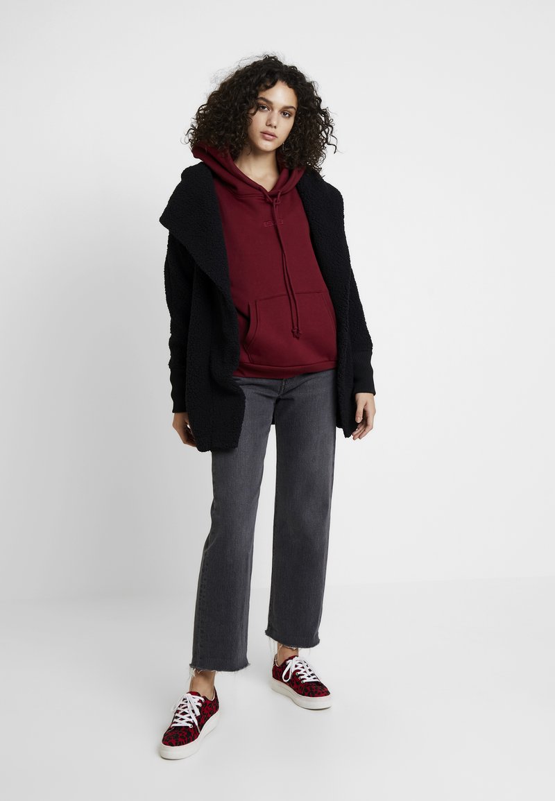 Schwarzer strukturierten Cardigan über einem bordeauxfarbenen Kapuzenpullover, kombiniert mit grauen weiten Jeans und roten Sneakern mit Leopardenmuster.