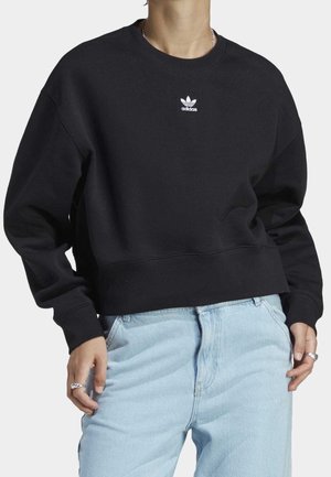 Personne portant un sweat-shirt Adidas noir avec un petit logo blanc sur la poitrine et un jean bleu clair, mains à côté des hanches.