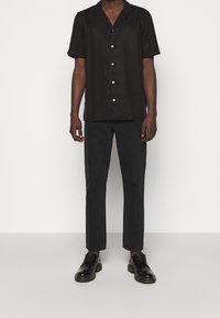 Chemise noire à manches courtes avec un col, boutons blancs, associée à un pantalon slim noir et des chaussures noires, se tenant sur un fond neutre.