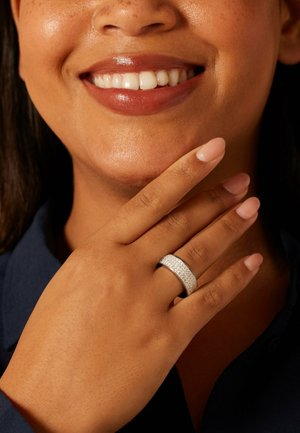 Zilveren ring met een getextureerde band en heldere ingelegde edelstenen. Hand van model met neutrale nagellak rustend op de wang, met een egale huidtint.