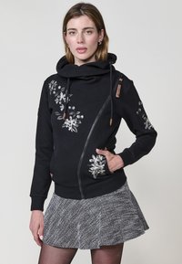 Sudadera negra con cuello de capucha, textura de espiga y bordado floral en el lado izquierdo. Cuenta con cierre de cremallera y bolsillos frontales.