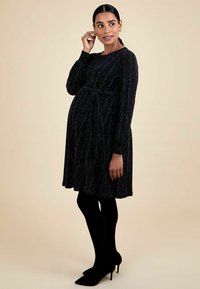 Vestido de maternidad negro con mangas largas, cintura fruncida, textura brillante y falda a la rodilla. Combinado con medias negras y tacones.