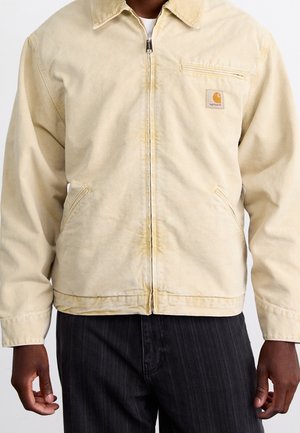 Mężczyzna w beżowej kurtce Carhartt na zamek z kołnierzem, przednią kieszenią na piersi i dwiema bocznymi kieszeniami, w zestawie z ciemnymi spodniami w prążki.
