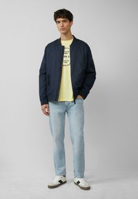 Marine bomberjack, gele graphic T-shirt, lichtblauwe jeans en witte sneakers met donkere accenten. Casual outfit gemaakt van lichtgewicht stof.