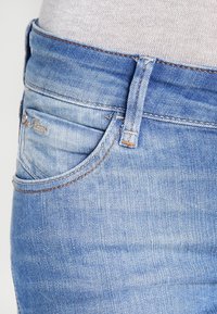 Světle modré džíny s klasickým střihem, které mají jemné vzory praní, oranžové prošití a malou nášivku na přední kapse.