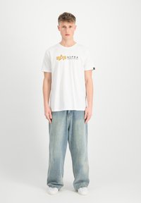 Bílý bavlněný tričko s černým a žlutým logem "ALPHA INDUSTRIES". V kombinaci s volnými světle modrými džíny a bílými teniskami.