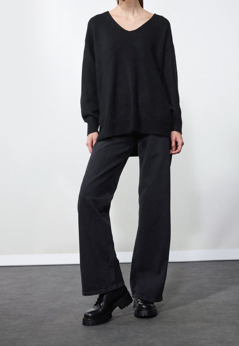 Personne portant un pull noir ample en V, un pantalon noir à jambes larges et des chaussures noires épaisses à lacets, debout sur un sol gris.