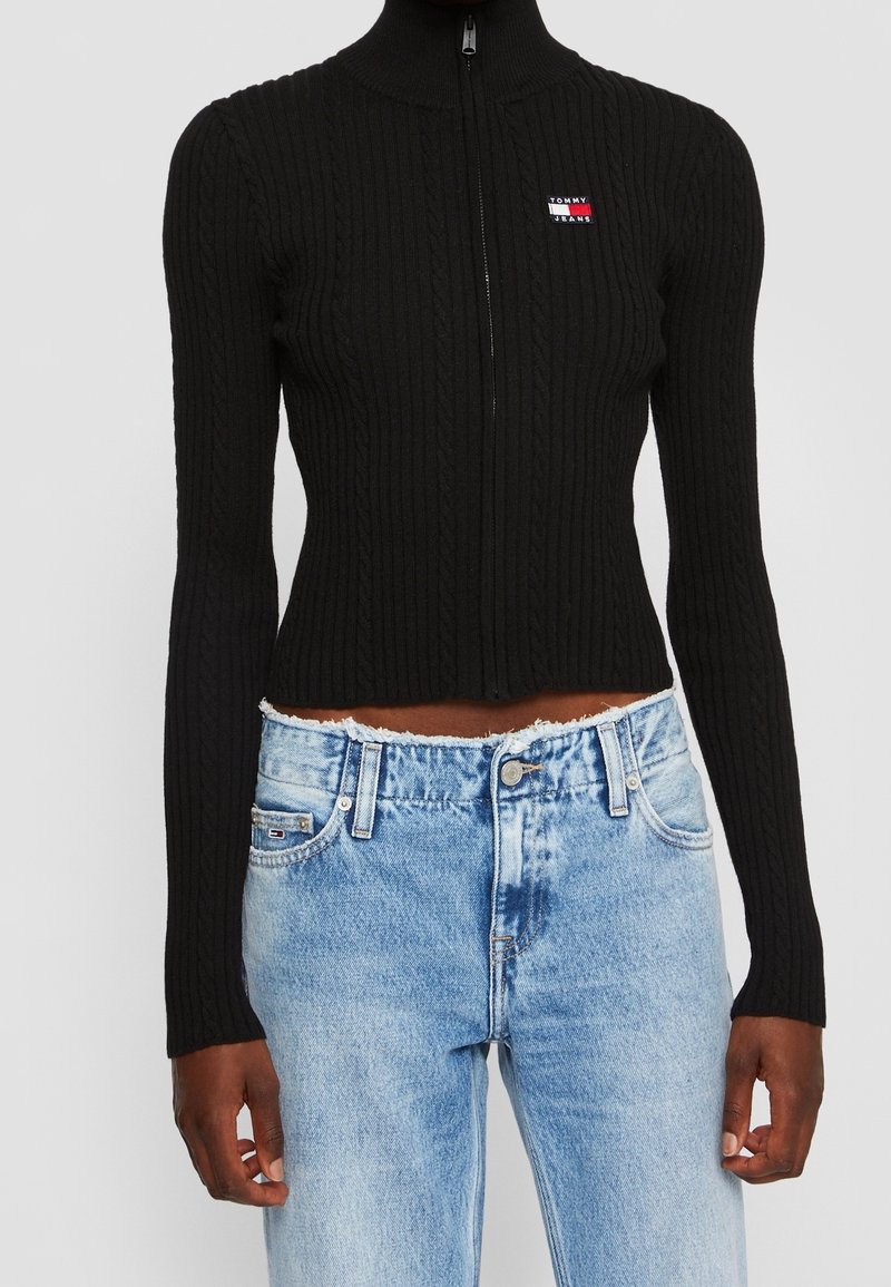 Pull à zip noir côtelé avec col montant, arborant un petit patch logo, associé à un jean en denim bleu clair déchiré.