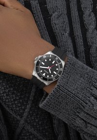 Csukló fekete gumiszíjas órával, amelyen 10:10 látható, rozsdamentes acél tok, fekete számlap, fehér mutatók, szürke kötött pulóver ujján viselve.