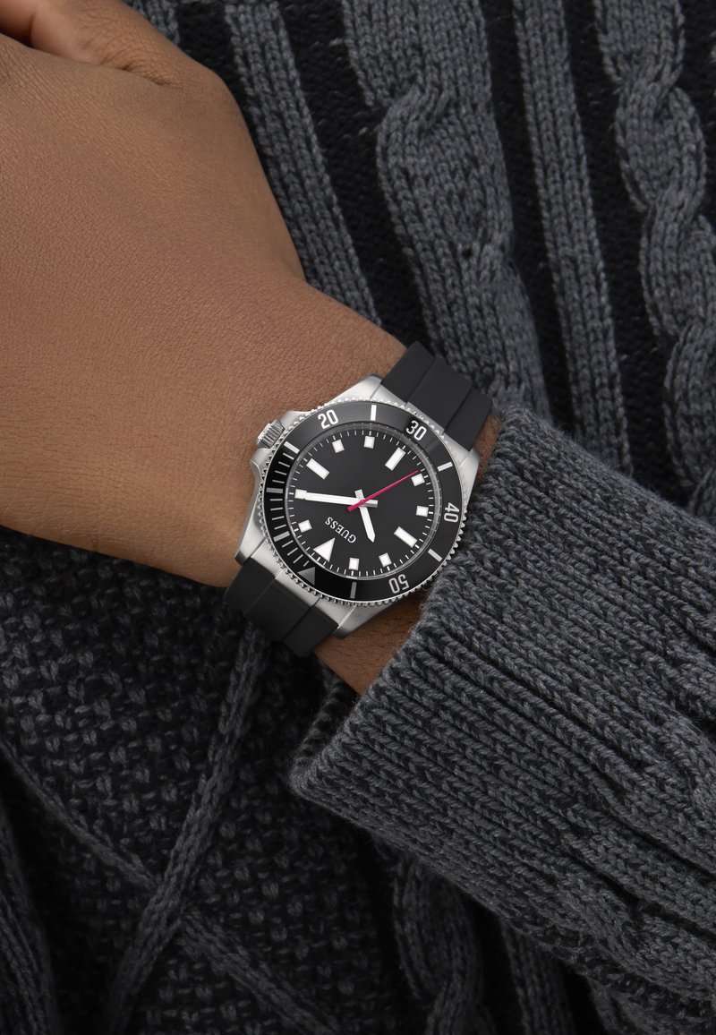 Csukló fekete gumiszíjas órával, amelyen 10:10 látható, rozsdamentes acél tok, fekete számlap, fehér mutatók, szürke kötött pulóver ujján viselve.