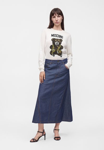 Femme portant un sweat-shirt blanc Moschino avec un motif ours en peluche, une longue jupe en jean bleue et des sandales noires à talons hauts, debout sur un fond blanc.