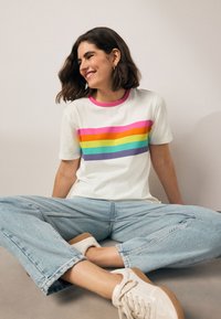 Fehér rövid ujjú póló több színű vízszintes csíkos mintával. Rózsaszín gallérral rendelkezik. Világoskék farmerrel és fehér sportcipővel párosítva.