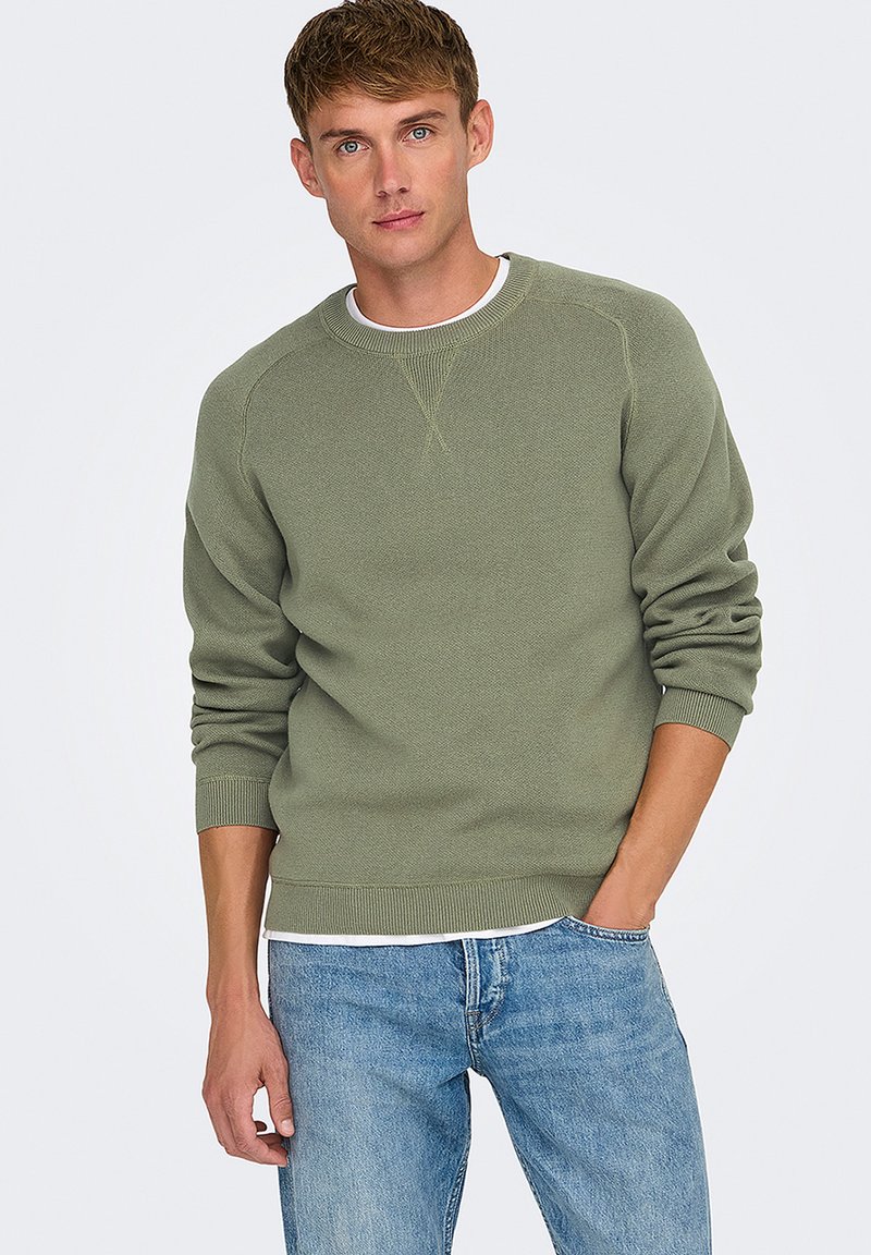 Maglione verde in tessuto con colletto a girocollo e polsini a costine, indossato sopra una t-shirt bianca, abbinato a jeans azzurri chiari.