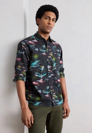 Homme debout contre un mur clair portant une chemise à boutons à imprimé abstrait multicolore avec les manches retroussées et un pantalon vert olive.