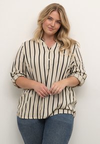 Beige overhemd met zwarte verticale strepen, met een V-hals en opgerolde mouwen, gecombineerd met blauwe spijkerbroeken.