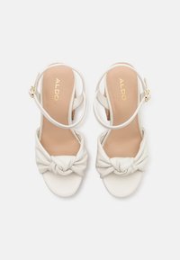 Sandales à talons blanches avec un design plissé noué, une bride de cheville réglable, et une semelle beige avec un marquage doré. Matière en cuir lisse.
