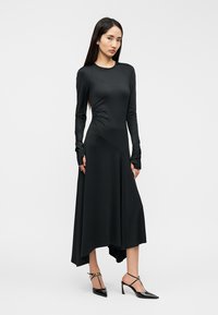 Μαύρο φόρεμα με μακριά μανίκια, στενή μπούστα και φουσκωτή ασύμμετρη φούστα. Ομαλή υφή, ραφές που διαμορφώνουν το σχήμα και μανσέτες με άνοιγμα για τα δάχτυλα.