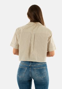 Camicia corta con righe verticali beige e marroni, maniche corte e dettaglio in yoke sul retro. Abbinata a jeans in denim blu a vita alta.