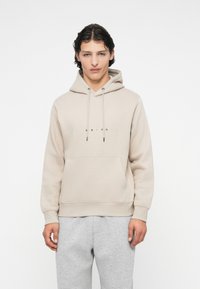Ung mand iført en beige hættebluse med snoretræk og kængurulomme, parret med lys grå sweatpants, stående mod en hvid baggrund.