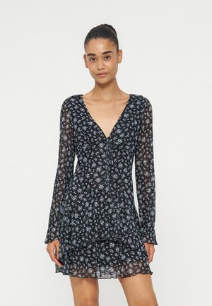 Jeune femme portant une robe fleurie noire transparente à manches longues et col en V profond, debout devant un fond blanc uni.