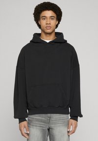 Melnā kapuce ar priekšējo kabatu, brīvs piegrieziens un ribveida aproces. Audums šķiet mīksts ar gludu tekstūru. Logotips diskrēti izvietots uz krūtīm.