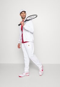Chaqueta atlética blanca y pantalones, camiseta roja debajo, gorra blanca, sosteniendo una raqueta de tenis, con zapatillas de tenis rosas y blancas.