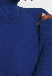 Sweter z dekoltem wystającym w głębokim niebieskim kolorze, o dopasowanym kroju, z prążkowanym dekoltem i długimi rękawami z wydłużonymi mankietami dla lepszej osłony.