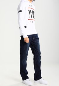 Bílá mikina s kapucí a grafickým designem v černých akcentem, kombinovaná s tmavě modrými džíny a bílými teniskami. Pohodlný vzhled a volný střih.