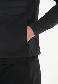 Sort jakke med en let polstret overdel, der har en lynlåslomme i siden og et glat, mat finish stof.
