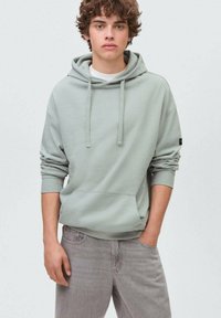 Ljusgrön huvtröja i mjukt tyg, med känguruficka, dragsko och ribbade muddar, i kombination med ljusgråjeans.