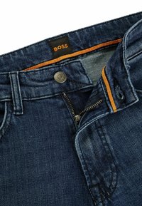 Mörkblå denimjeans med en öppen dragkedja, metallknapp märkt "BOSS" och orange kant längs insidan av midjebandet.