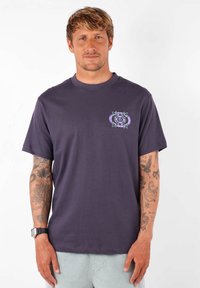 T-shirt en coton violet à manches courtes avec un motif floral sur le côté gauche de la poitrine. Présenté sur un modèle tatoué.