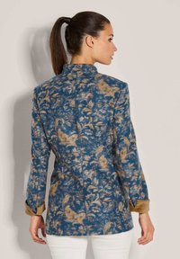 Vrouw met een paardenstaart die een blauw en beige bloemenpatroon jasje met opgerolde mouwen en witte broek draagt, met de rug naar de camera gekeerd tegen een eenvoudige achtergrond.