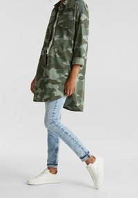 Persona vestida con una camisa larga de camuflaje verde, jeans ajustados de un azul claro y zapatillas blancas, de pie con una pierna cruzada sobre la otra.