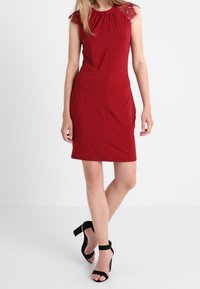 Femme portant une robe rouge sans manches à mi-cuisses avec des épaules en dentelle et des sandales noires à talons hauts, se tenant les jambes croisées.