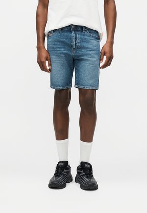Homme portant un short en denim bleu moyen, des chaussettes blanches montantes, des baskets Diesel noires et un t-shirt blanc sur un fond blanc uni.