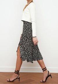 Cropped top bianco a maniche lunghe abbinato a una gonna midi nera con motivo floreale dotata di uno spacco laterale, completato da sandali neri con tacco a fascia.
