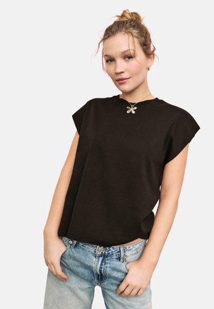 Frau mit blondem Haar zu einem Dutt gebunden, trägt ein schwarzes ärmelloses Oberteil, hellblaue Jeans und eine blumenförmige Halskette, posiert mit den Händen in den Taschen.